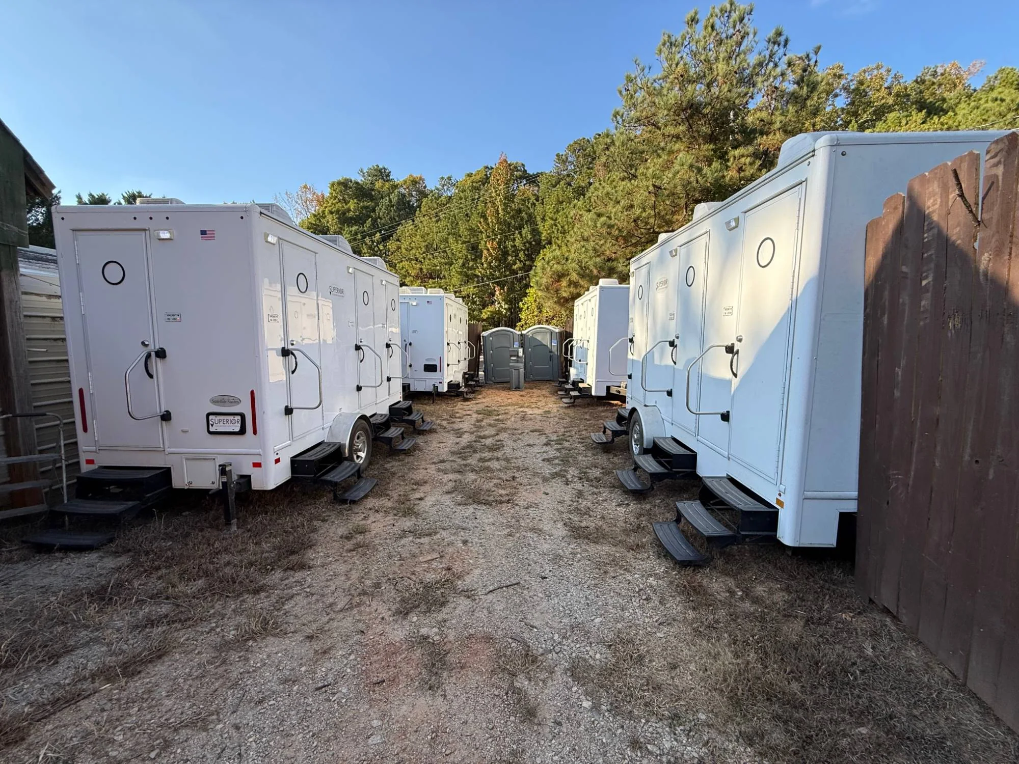 Two rows of Superior Location Solutions restroom trailers facing each other across a dirt service lane at the Georgia Renaissance Festival, with portable toilets at the far end